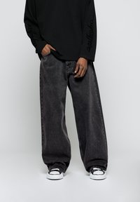 Camisa negra de manga larga con texto en los puños, combinada con unos pantalones vaqueros oscuros y holgados de color gris oscuro y zapatillas en blanco y negro.