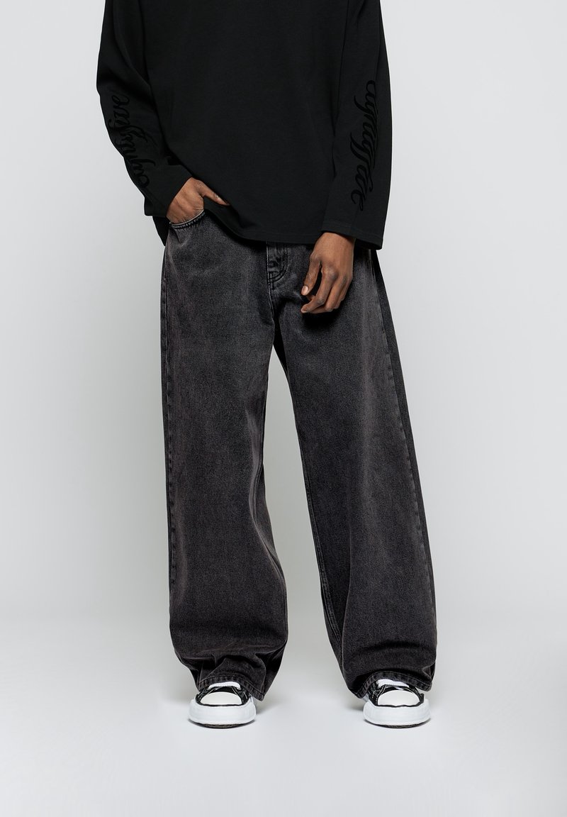 Camisa negra de manga larga con texto en los puños, combinada con unos pantalones vaqueros oscuros y holgados de color gris oscuro y zapatillas en blanco y negro.
