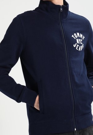 Námořnicky modrá bunda na zip s vysokým límcem, bílým logem "Tommy NYC Jeans" na levé straně hrudi a rukama v bočních kapsách.