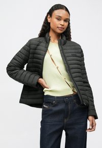 O femeie tânără cu păr creț poartă o jachetă neagră matlasată peste un top cu butoni de culoare galben pal și blugi de culoare albastru închis, pozând pe un fundal alb.