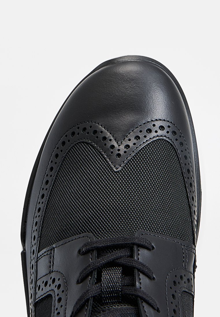 Des chaussures en cuir noir avec une tige en maille texturée, des perforations décoratives et un motif en forme de cœur au niveau des orteils. Lacets noirs inclus.