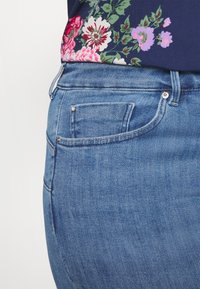 Jeans de denim azul com uma visão detalhada, apresentando uma cintura média, bolso traseiro com um botão decorativo e uma textura subtil. Parte de uma blusa floral visível.