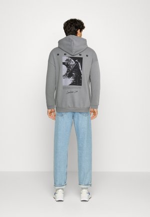 Personne portant un sweat à capuche gris avec un motif noir et blanc et du texte dans le dos, un jean bleu clair et des baskets blanches, debout sur un fond blanc uni.