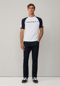 Biely tričko s námorníčko modrými raglánovými rukávmi a predným logom "HACKETT". Nosené s tmavomodrými riflami a námorníčko modrými teniskami s bielymi akcentmi.