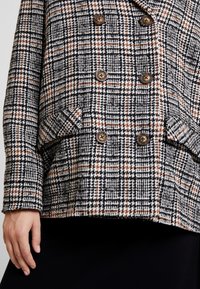 Dvojřadový blejzr s černobílým houndstooth vzorem a hnědými prvky. Má strukturovanou tkaninu a hnědé knoflíky, dvě postranní kapsy.