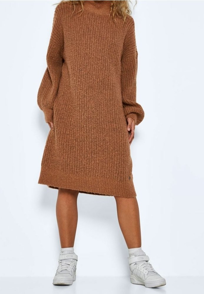 Femme portant une robe midi marron en maille à manches longues, associée à des baskets blanches montantes, debout devant un fond uni.