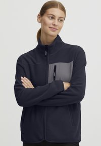 Fleece jacket in navy blue, featuring a gray chest pocket with zip, high collar, and side pockets. Soft texture and relaxed fit.