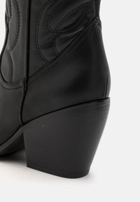 Stivaletto in pelle nera con superficie texturizzata, dettagli cuciti e tacco nero a forma di cono. Il design presenta delicati motivi impressi.