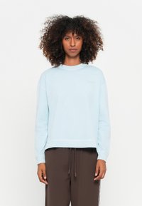 Felpa corta azzurro chiaro realizzata in tessuto morbido, con collo rotondo e spalle leggermente cadenti, abbinata a pantaloni jogger marrone scuro.