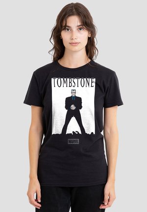 Young woman wearing a black T-shirt featuring Marvel's Tombstone character standing with clenched fists under the title "TOMBSTONE."