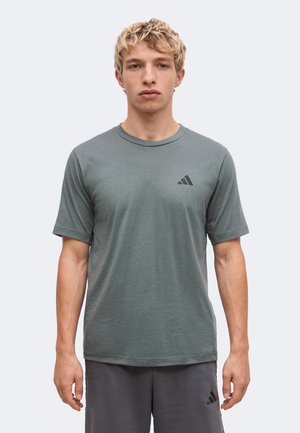 Млад мъж с къса руса коса, облечен в сива тениска Adidas и съответстващи сиви къси панталони, стои пред обикновен светъл фон.