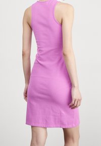 Žena v bezramenných, žebrovaných, šatových růžových šatech po kolena, zobrazena zezadu na jednoduchém světlém pozadí.