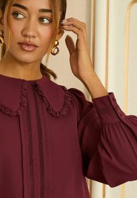 Vinröd blus med krage kantad med spets, veckade puffärmar och vertikala detaljer framtill, tillverkad av ett mjukt tyg.