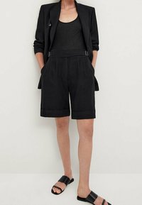 Zwarte blazer van linnenmix over een zwart tanktop, gecombineerd met op maat gemaakte zwarte shorts met omgeslagen zoom en zijzakken. Zwarte slip-sandalen.
