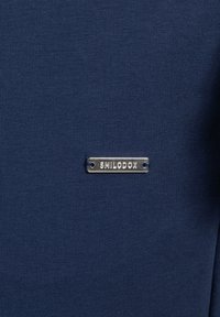 Námořnická modrá, vroubkovaná tkanina se stříbrným kovovým štítkem, na kterém je nápis "SMILODOX", se dvěma malými dírkami pro připevnění.