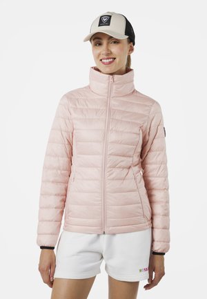 Chaqueta acolchada de color rosa claro con un alto collar, textura acolchada, cierre frontal de cremallera y bolsillos laterales. Lleva puesta con pantalones cortos blancos y un sombrero.