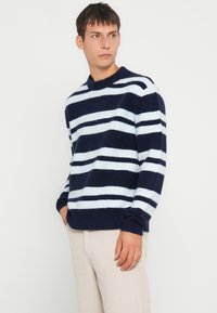 Maglione a righe blu navy e azzurre in un punto di maglia testurizzato. Presenta un colletto rotondo e polsini e orlo a costine. Indossato con pantaloni chiari.