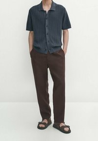 Homme portant une chemise en maille à manches courtes gris foncé boutonnée, un pantalon marron foncé et des sandales marron foncé à bout ouvert avec des semelles noires. Mains dans les poches.