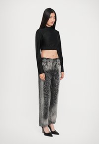 Woman wearing long-sleeve black cropped top and high-waisted gray jeans with black spotted pattern, paired with black high heels, standing sideways.