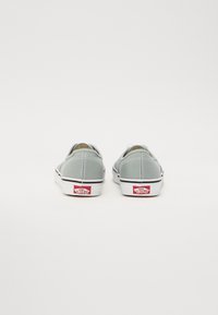 Grå canvas slip-ons med en vit gummisula, svarta detaljer och röd logotyp på hälen, som har en enkel och strömlinjeformad form.
