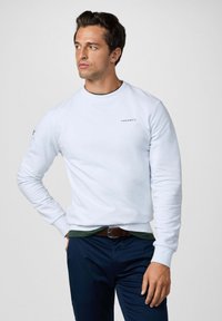 Sudadera blanca con cuello redondo y puños acanalados, que presenta un pequeño logo "HACKETT" en el pecho. Combinada con pantalones azul marino y una camisa verde debajo.