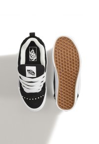 Zapatillas de ante negras con cordones blancos, que presentan tachuelas plateadas en la puntera. La suela es blanca con un patrón de diamantes y fondo de goma marrón.