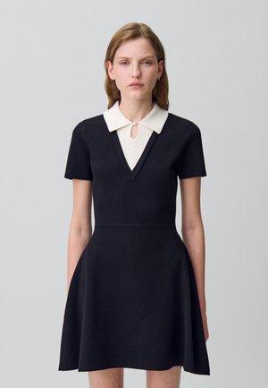 Abito nero a maniche corte con scollatura a V, caratterizzato da un colletto bianco e un motivo testurizzato sul corpetto. Gonna svasata e vita aderente.