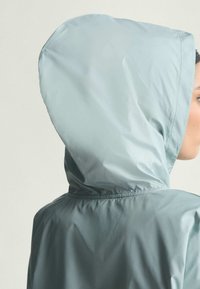 Letvægts hættejakke i lys blå stof med en glat overflade, der har en elastisk talje og et fleksibelt hætte-design.