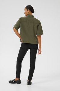 Chemise olive à manches courtes avec une coupe droite, associée à un jean slim noir et des mocassins noirs, vue de dos.