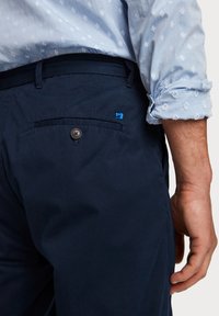 Námořnické modré kalhoty s elegantním designem, obsahující zadní kapsu, hnědý knoflík a malý modrý logo akcent. Měkká textilie.