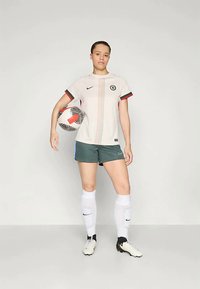 Tenue de football Nike comprenant un maillot crème clair avec des accents rouges et verts, associé à un short vert foncé, des chaussettes blanches et un ballon noir et blanc.