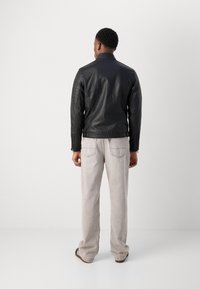 Jachetă din piele neagră cu guler în picioare, detalii ribbate pe umeri și manșete cu fermoar, asociată cu blugi gri deschis și sandale negre tip slide.