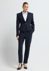 Námořnický elegantní oblek s přiléhavým blejzrem a úzkými kalhotami, doplněný bílou košilí. Texturované černé vysoké podpatky dokončují vzhled.