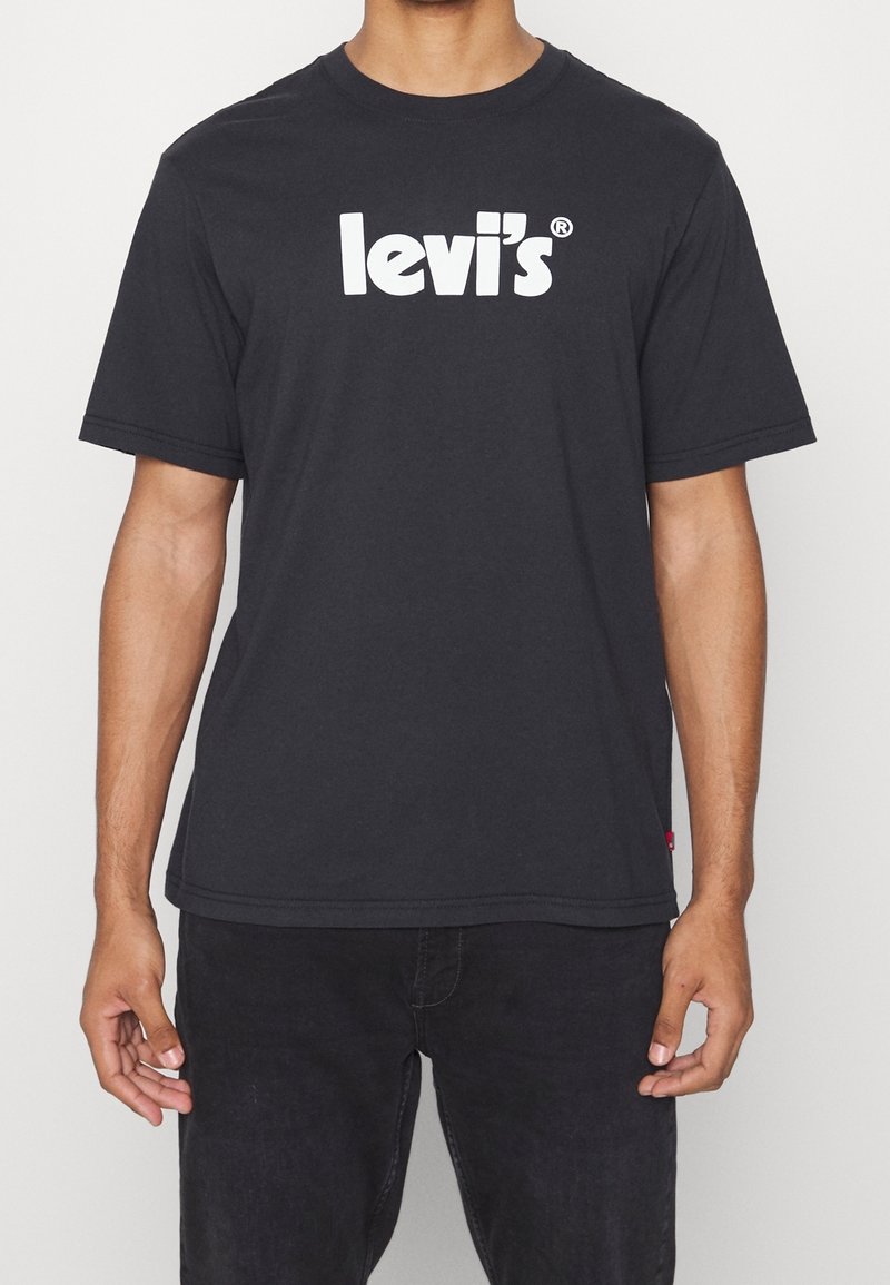 Homme portant un t-shirt noir Levi's avec du texte blanc en logo sur la poitrine et un pantalon noir, se tenant devant un fond uni.
