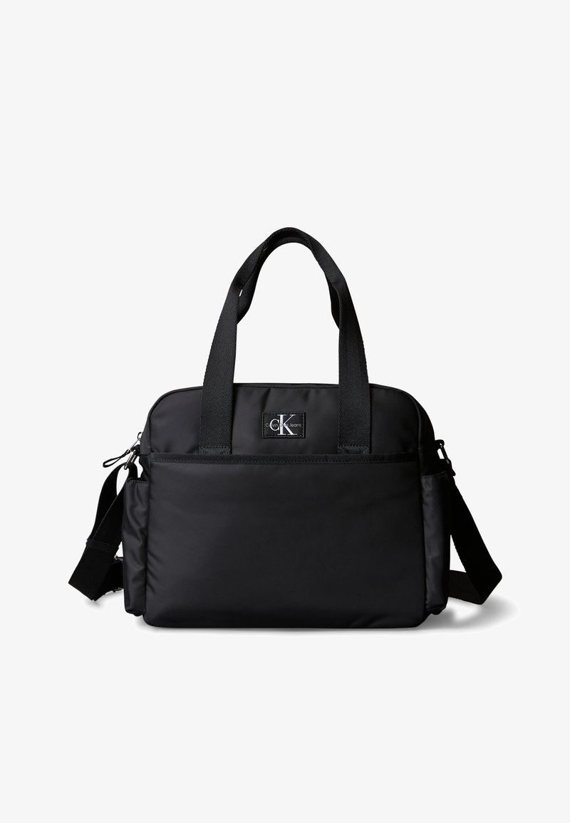 Μαύρη τσάντα ώμου και χειρός Calvin Klein με μπροστινή τσέπη, δύο χερούλια και ρυθμιζόμενο λουράκι σε λευκό φόντο.