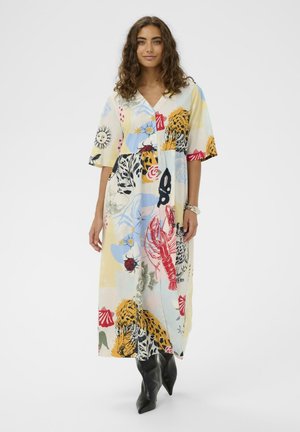 Kobieta w kolorowej sukience midi z motywami zwierzęcymi i przyrodniczymi, w parze z czarnymi spiczastymi botkami, stojąca na białym tle.