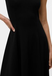 Robe noire avec un corsage ajusté, une jupe évasée et une texture lisse. La main du modèle repose sur la couture latérale. Design simple et élégant.