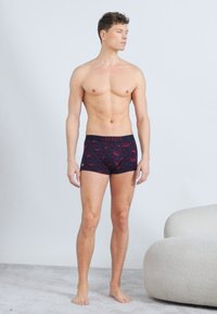 Άνδρας χωρίς πουκάμισο, ξυπόλητος σε χαλί, φοράει μαύρα και κόκκινα boxer brief με σχέδια της Lacoste, δίπλα σε μια λευκή στρογγυλεμένη πολυθρόνα.