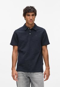 Granatowa koszulka polo z teksturowanej tkaniny, krótki rękaw i kołnierzyk. Posiada dwie guziki i małe logo na piersi.