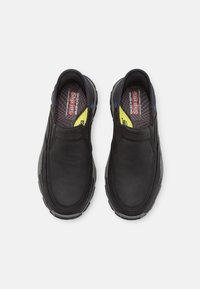 Chaussures noires à enfiler avec un dessus en cuir lisse, une semelle intérieure texturée noire et des semelles en caoutchouc grises. Elles présentent des accents bleus et jaunes.