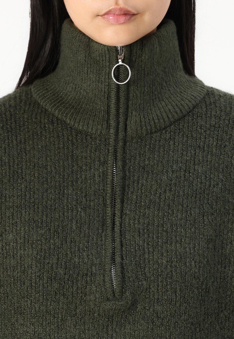 Dunkelgrüner Strickpullover mit hohem Kragen, ausgestattet mit einem silbernen Reißverschluss auf der Vorderseite, der einen strukturierten Stoff präsentiert.