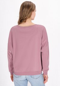Př oversized dlouhý rukáv svetr v světlé mauvé, s kulatým výstřihem a pletenými manžetami, v kombinaci se světlými modrými džíny.
