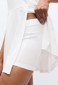 Λευκό αθλητικό τοπ με πλαϊνή σχισμή, που αποκαλύπτει λευκό εφαρμοστό σορτς από κάτω. Ελαφρύ, λείο ύφασμα με ελαστική μέση και καθαρές λεπτομέρειες ραφής.