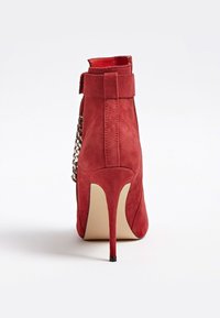 Bottine à talon aigu en daim rouge avec un détail chaîne sur le côté et un talon haut fin sur un fond blanc uni.