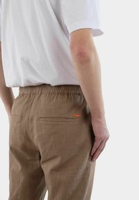 Bruna linbyxor med elastisk midja, två bakfickor och en orange logodetalj. Bärs tillsammans med en vit kortärmad skjorta.