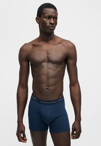 Άνδρας μοντέλο με σκουρόχρωμο δέρμα που φοράει σκούρα μπλε μπόξερ Tommy Hilfiger, στέκεται μπροστά σε απλό ανοιχτό γκρι φόντο.