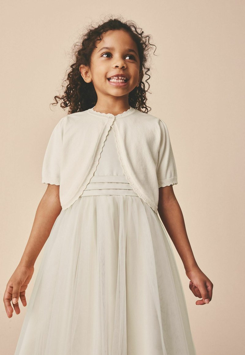 Jeune fille souriante aux cheveux bouclés portant une robe blanche et un cardigan blanc à manches courtes, debout devant un fond beige.