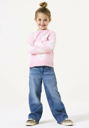 Rosa Pullover mit weißem Blumenmuster, lockere blaue Jeans und gemusterte Sneakers. Die Haare zu einem Dutt frisiert. Steht mit verschränkten Armen.