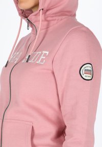 Felpa rosa con zip realizzata in tessuto morbido, dotata di testo argentato sul petto, cappuccio regolabile e una toppa con logo sulla manica sinistra.