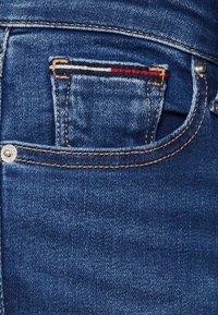 Jeans de mezclilla azul oscuro con un pequeño bolsillo frontal, costuras visibles y un remache de metal en el lateral. Textura suave con un ligero desvanecimiento.